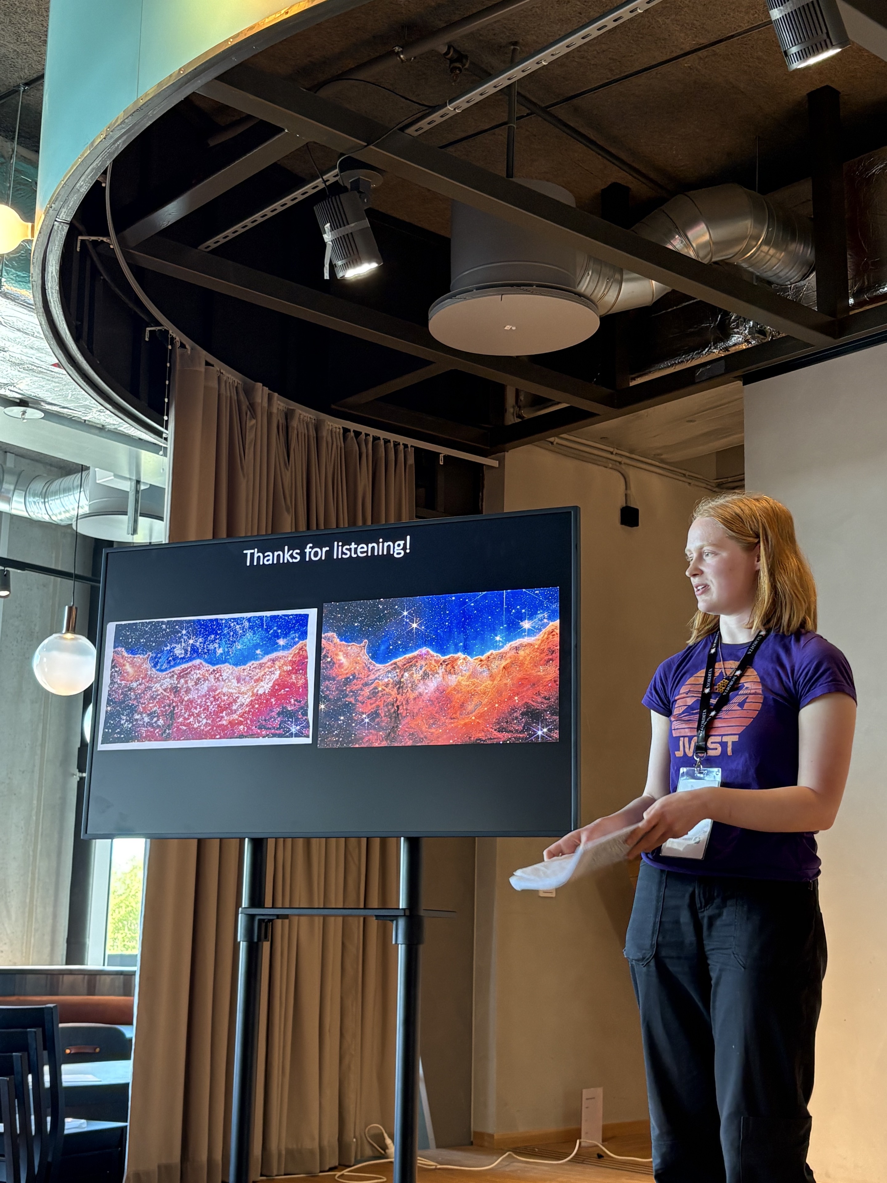 Harriet Wulff stands beside a presentation screen showing her Cosmic Cross Stitch and its James Webb Space Telescope inspiration image, the Carina Nebula's Cosmic Cliffs.